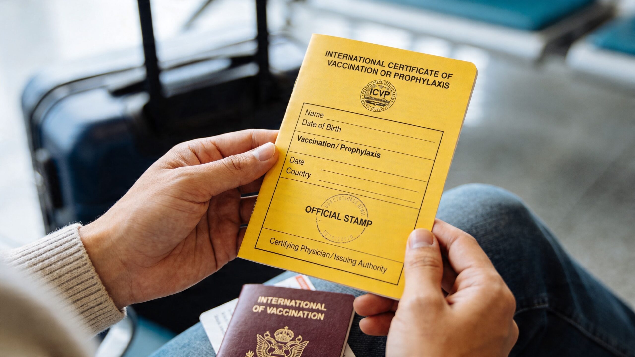 A person holds a yellow International Certificate of Vaccination or Prophylaxis booklet while sitting with luggage.