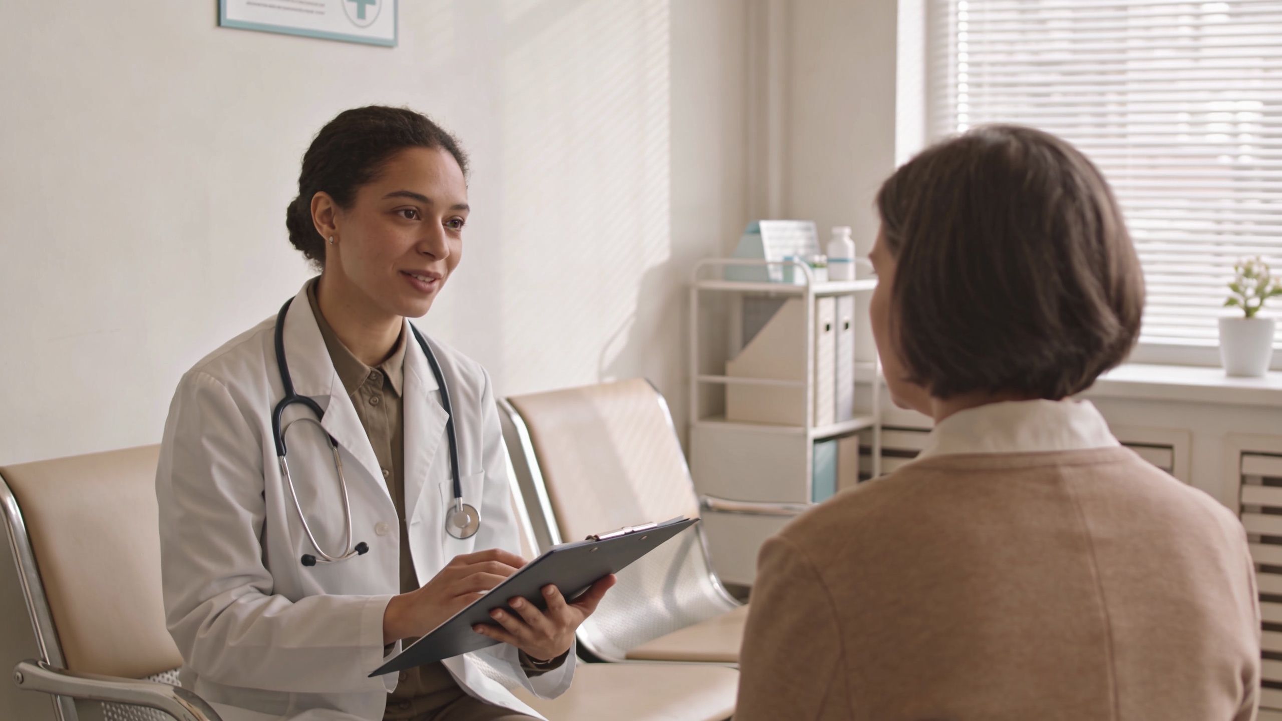 A female doctor in a white coat talks to a patient while holding a clipboard in an office.